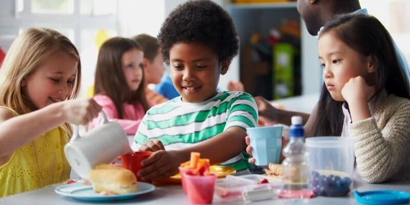 a girl in a yellow shirt pours water for a boy and another girl at her lunch table front of house software for school nutrition programs